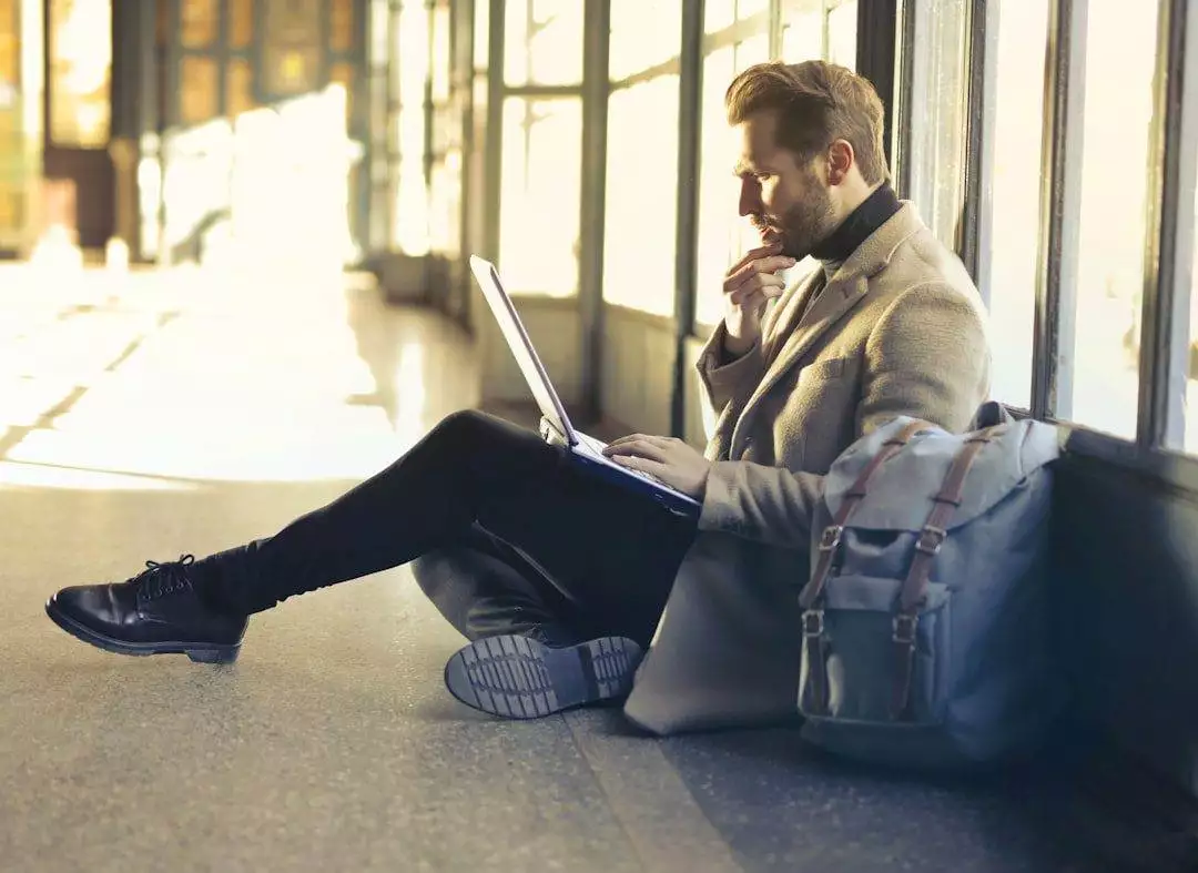 A man sitting on the floor by large windows, using a laptop on his lap, with a backpack beside him—perhaps researching a professional website design company or looking for a WordPress web designer for his small business.