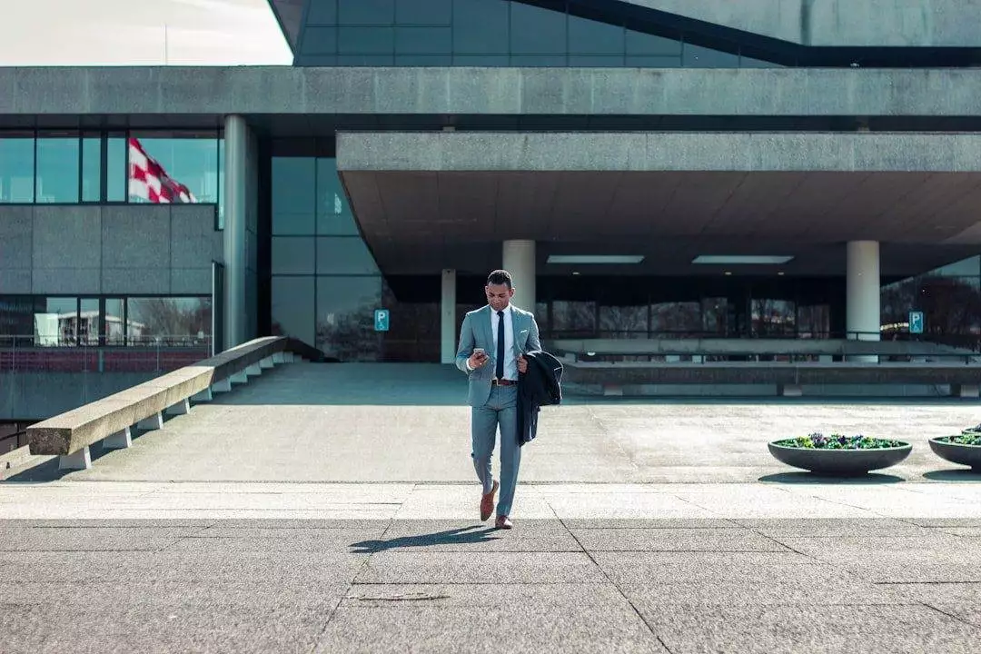 Unlocking Website Maintenance Success Metrics A man in a suit walks outside a modern building, holding a phone and jacket—perhaps discussing affordable web design services. A flag and large planters are visible in the background.