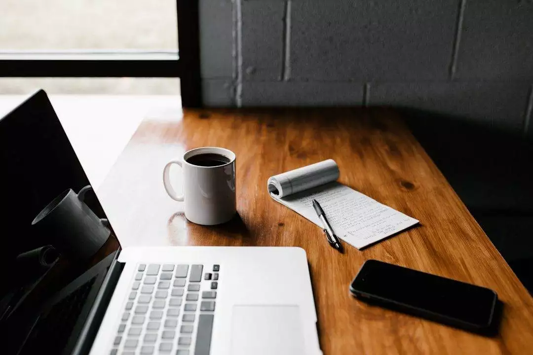 A wooden desk with a laptop, cup of coffee, notepad with handwritten notes, pen, and smartphone near a window—inspiring setup for any WordPress web designer working on creative projects.