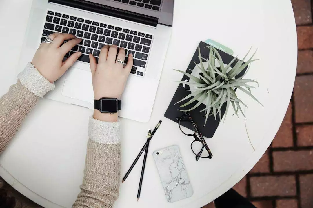 Person typing on a laptop at a round white table with a notebook, plant, smartphone, glasses, and pencils—perfect setting for a WordPress web designer offering affordable web design services.