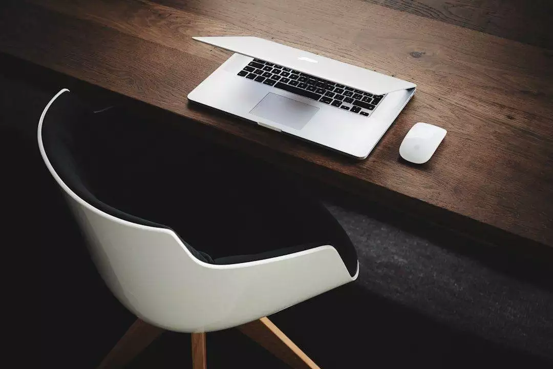 A partially closed laptop, white wireless mouse, and modern white chair on a dark wooden desk—perfect inspiration for small business website design.