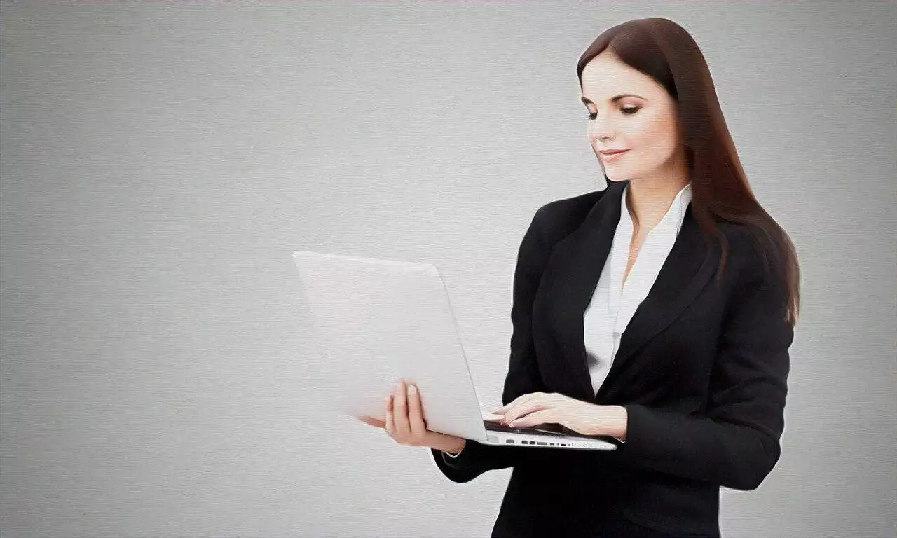 A woman in a black suit stands holding and typing on an open laptop against a plain light grey background, representing a professional website design company.