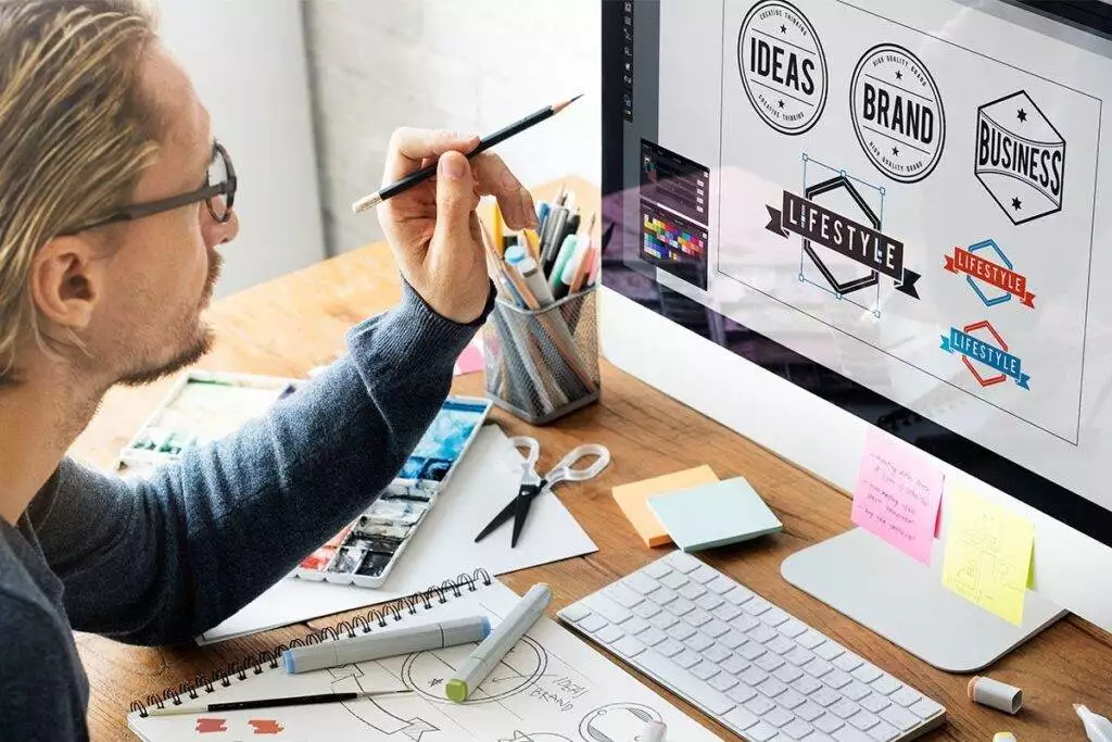 A person sits at a desk working on logo designs for a small business website design at a computer, surrounded by sketches, notes, and stationery items.