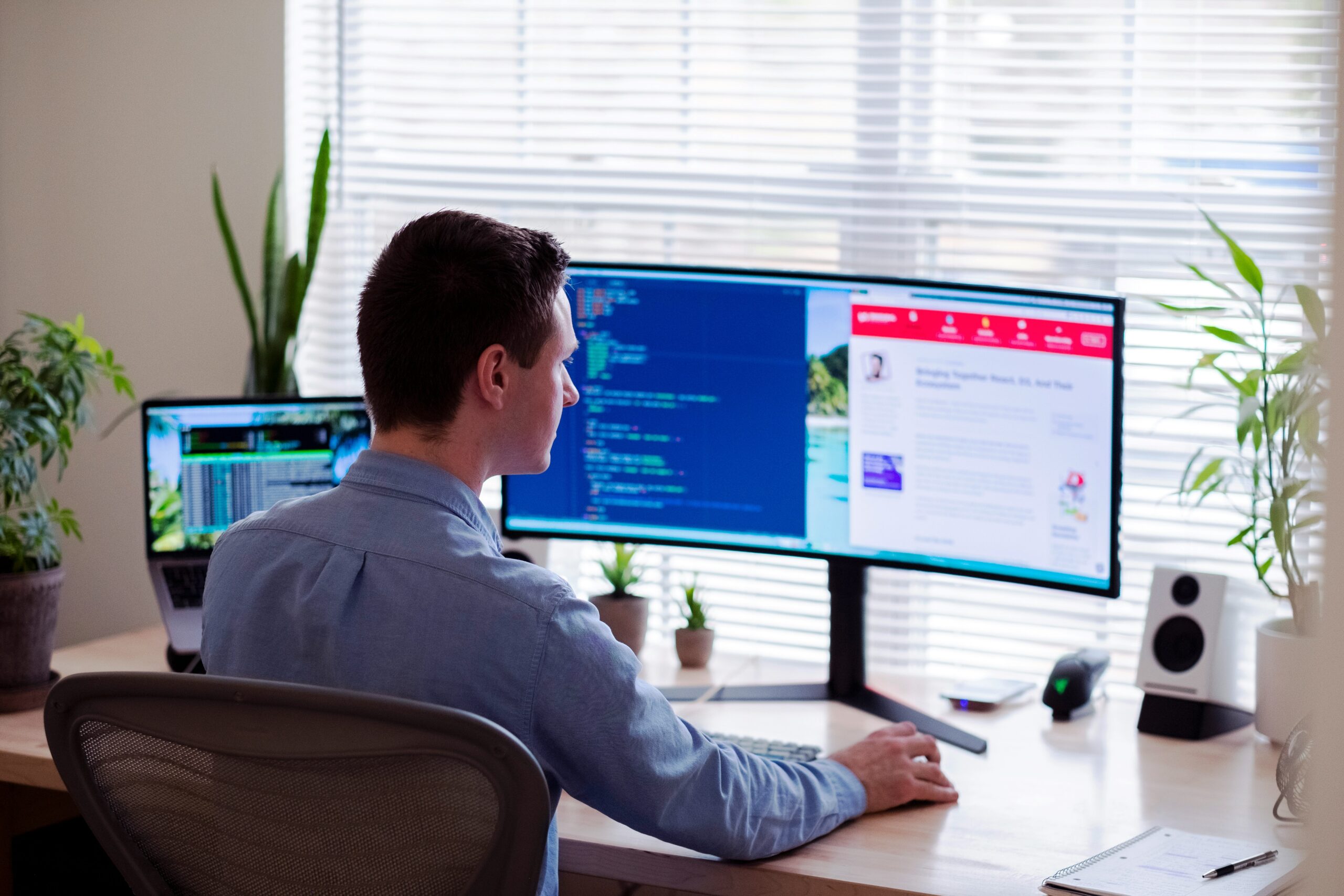 The Ultimate Guide to Web Development A person sits at a desk with a laptop and an ultra-wide monitor displaying code and a news website, surrounded by plants and office supplies.