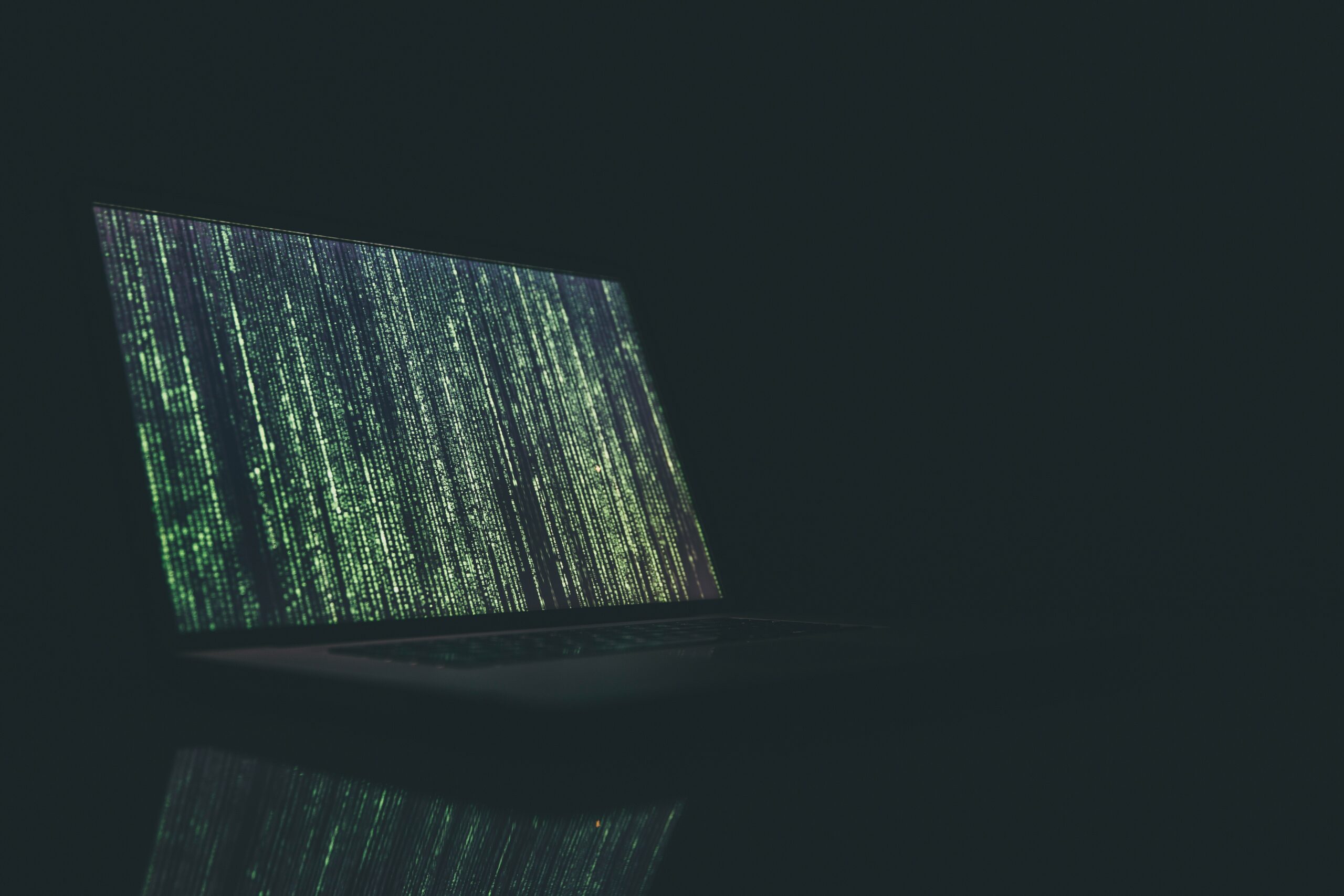 A laptop displaying green vertical lines of code on a dark screen, reminiscent of a digital or hacking interface, with a dark background.