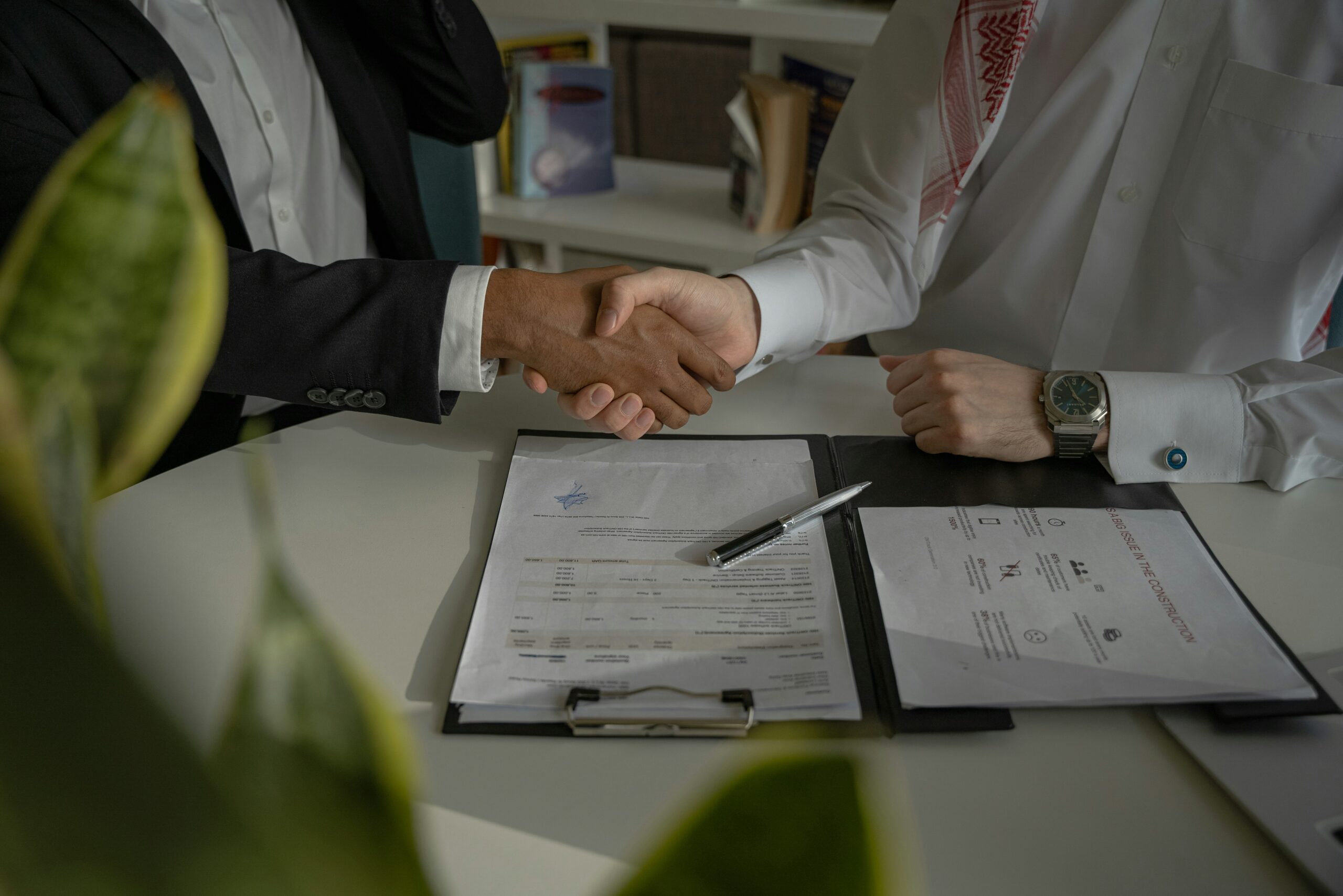 Why You Should Partner With a Dedicated Web Agency Two people shake hands across a desk with documents, a pen, and a clipboard visible, indicating a business agreement or contract signing.