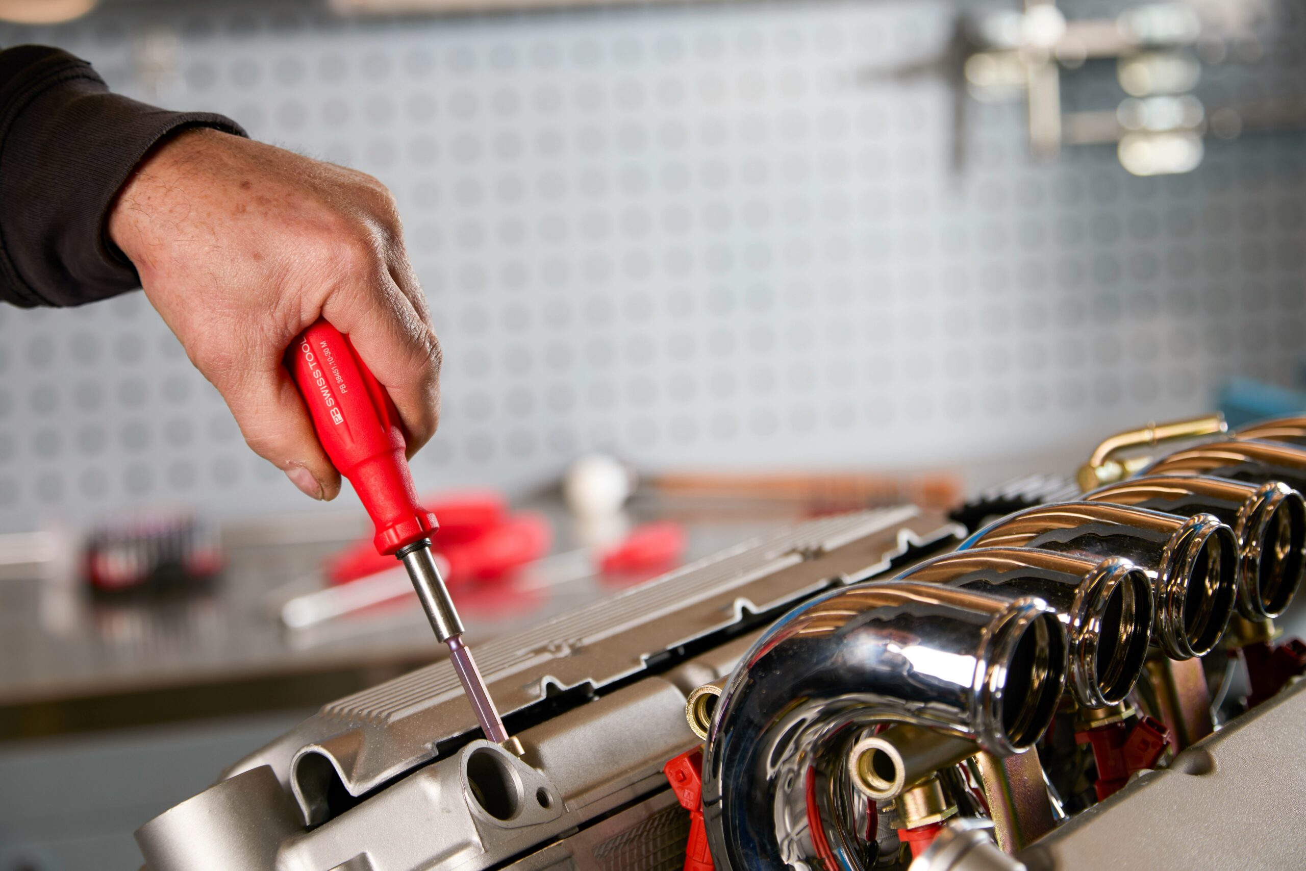A person uses a red-handled screwdriver to work on a metallic engine part in a workshop.