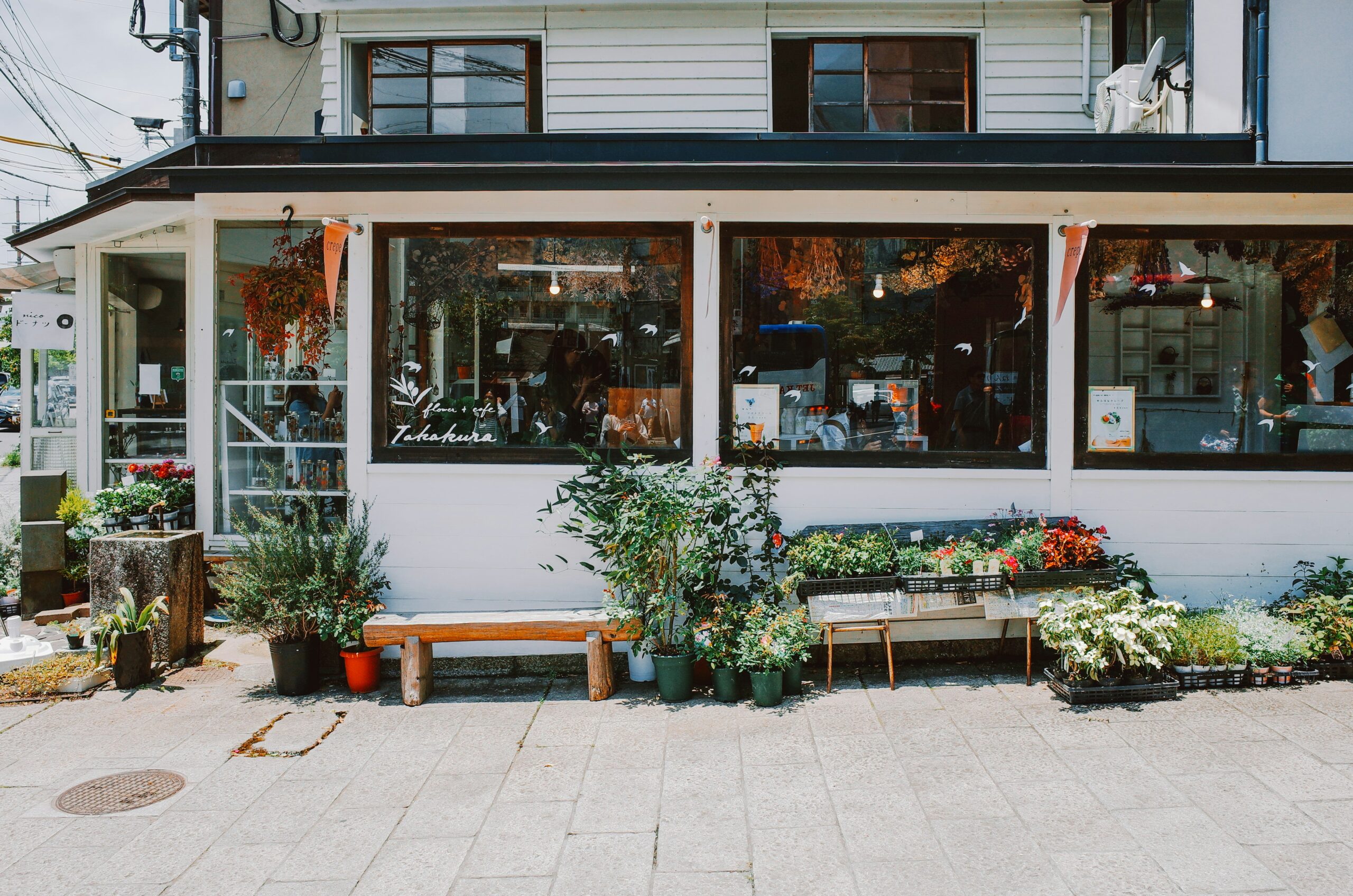 A small storefront with large windows displays various potted plants, flowers, and greenery arranged along the sidewalk in front.