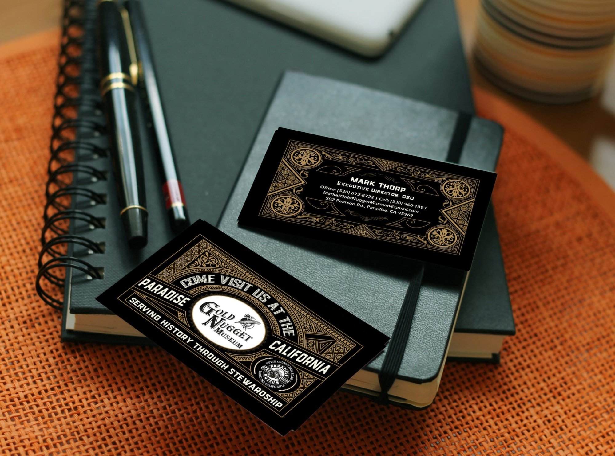 Portfolio Two business cards labeled “Gold Nugget Museum” rest on a black notebook with a pen and orange textured placemat underneath, reflecting the professionalism you’d expect from affordable web design services for small businesses.All-inclusive Website Design Services