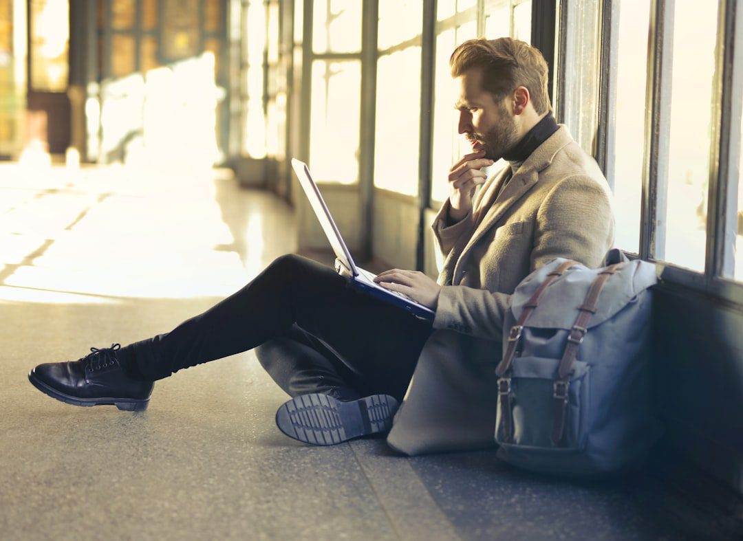 Unlock Your Online Potential with Custom Website Design A man sits on the floor by large windows, using his laptop with a backpack beside him. He appears focused, perhaps exploring affordable web design services or considering website maintenance and support for his next project.
