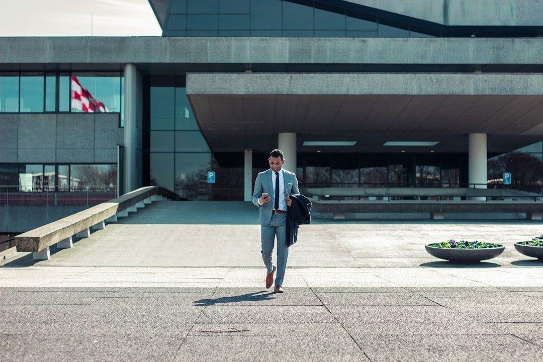 A man in a suit walks outside a modern building, holding a phone and jacket—perhaps discussing affordable web design services. A flag and large planters are visible in the background.