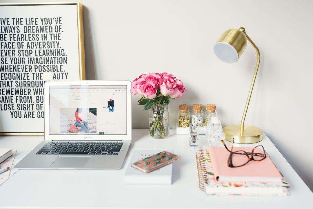 A tidy desk with a laptop, pink flowers in a vase, and a gold lamp sets the scene for an inspired web designer near me—complete with notebooks, glasses, phone, small jars, and a framed quote in the background.