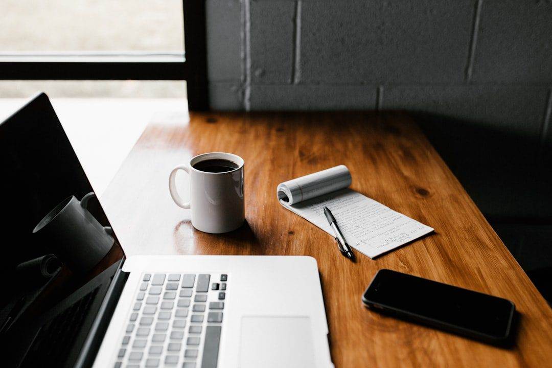 A wooden desk with a laptop, cup of coffee, notepad with handwritten notes, pen, and smartphone near a window—inspiring setup for any WordPress web designer working on creative projects.