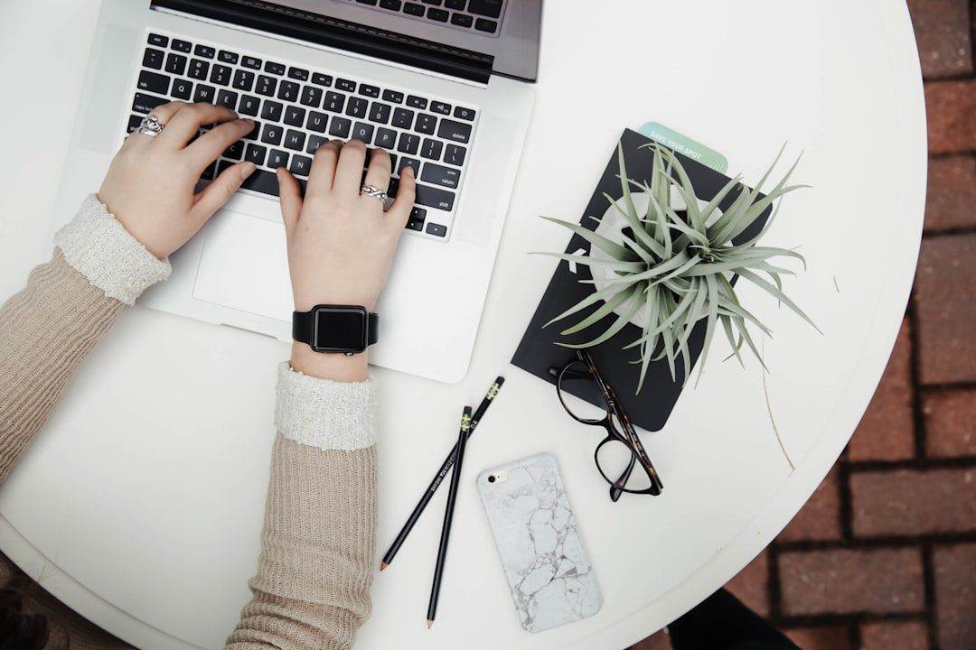 Person typing on a laptop at a round white table with a notebook, plant, smartphone, glasses, and pencils—perfect setting for a WordPress web designer offering affordable web design services.