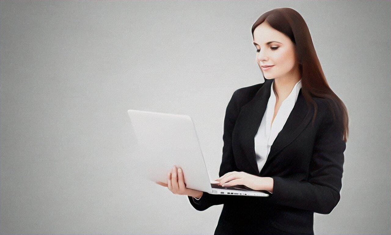 A woman in a black suit stands holding and typing on an open laptop against a plain light grey background, representing a professional website design company.