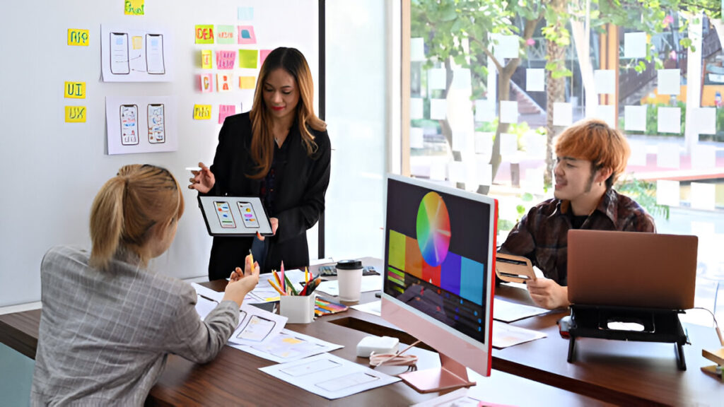 Three people in an office discuss mobile app designs using tablets, a desktop computer, and notes on a whiteboard—just like a professional website design company collaborating on small business website design projects.