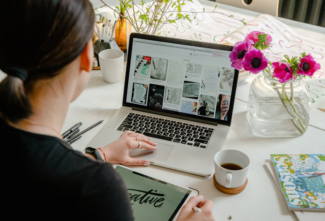 A person sits at a desk with a laptop displaying sketch designs for small business website design, a tablet showing "creative," a notebook, flowers in a vase, and a cup of coffee.