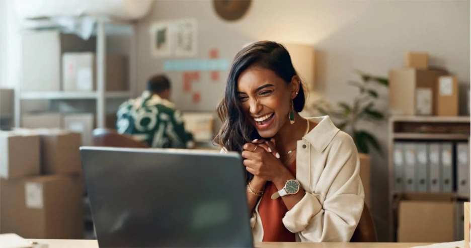 Content Marketing: Creating Engaging Content That Converts A woman sits at a desk, smiling and clasping her hands as she looks at a laptop screen, surrounded by storage boxes and another person working in the background—an inspiring scene of engaging content in action.