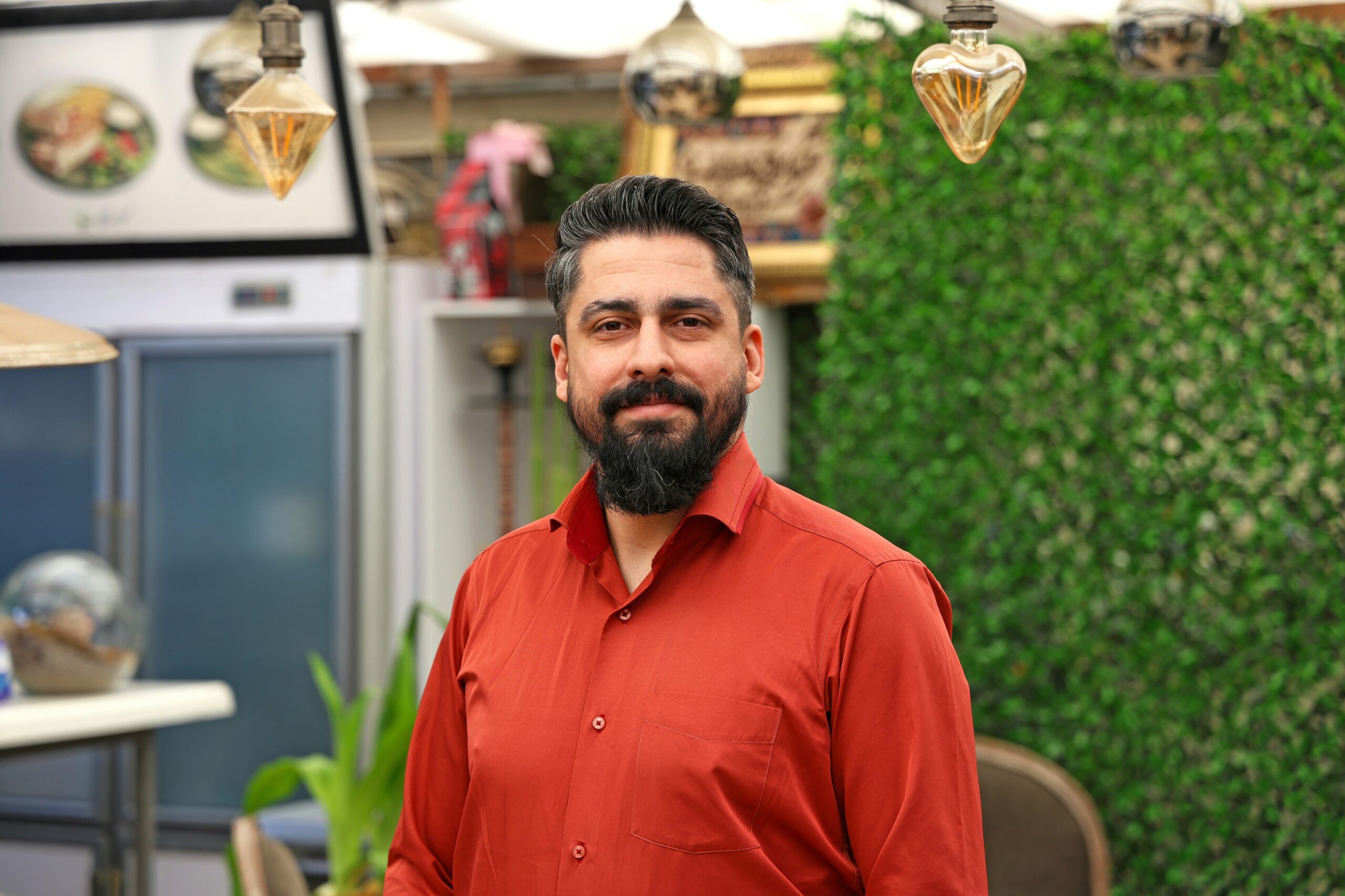 A man with a beard and mustache, wearing a red shirt, stands indoors in front of a green leafy wall and restaurant decor.