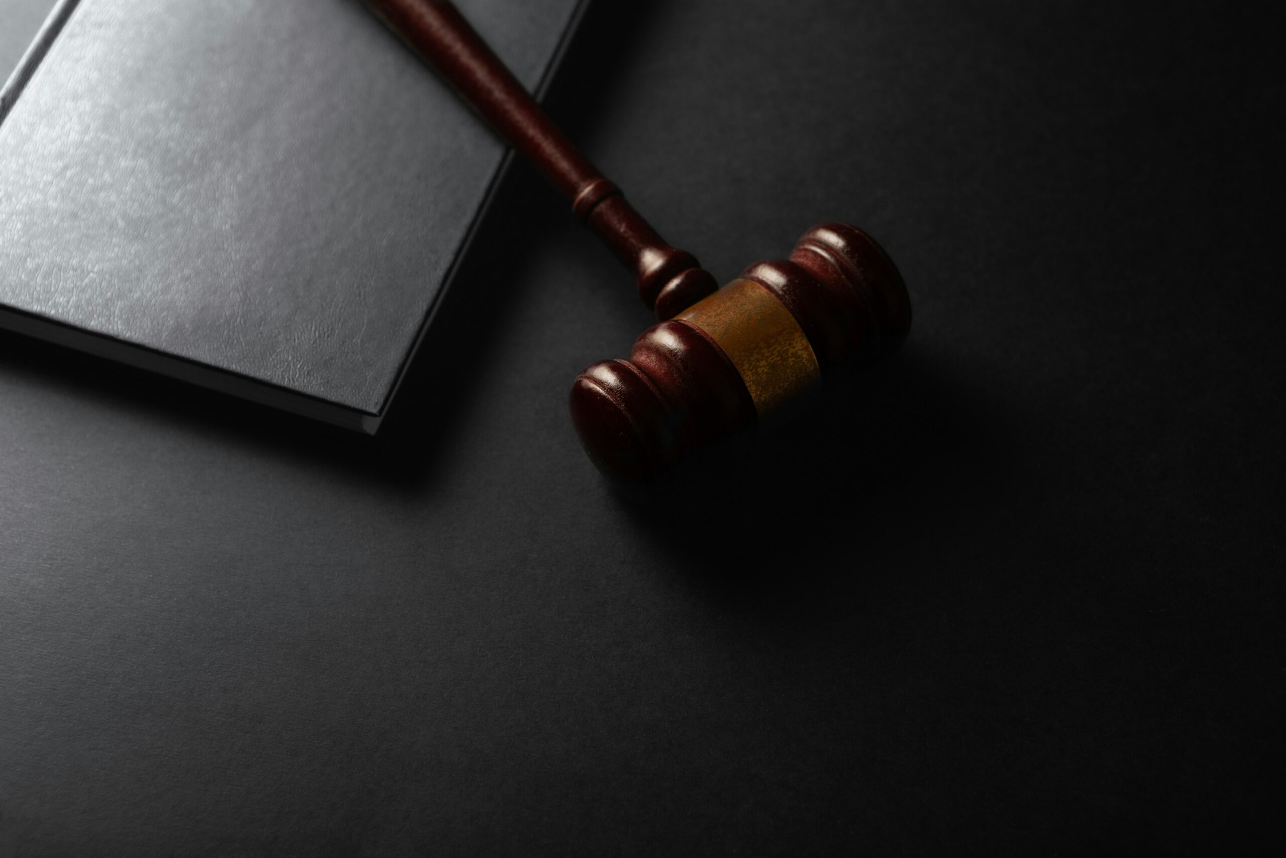 A wooden judge’s gavel rests next to a closed black notebook on a dark surface.