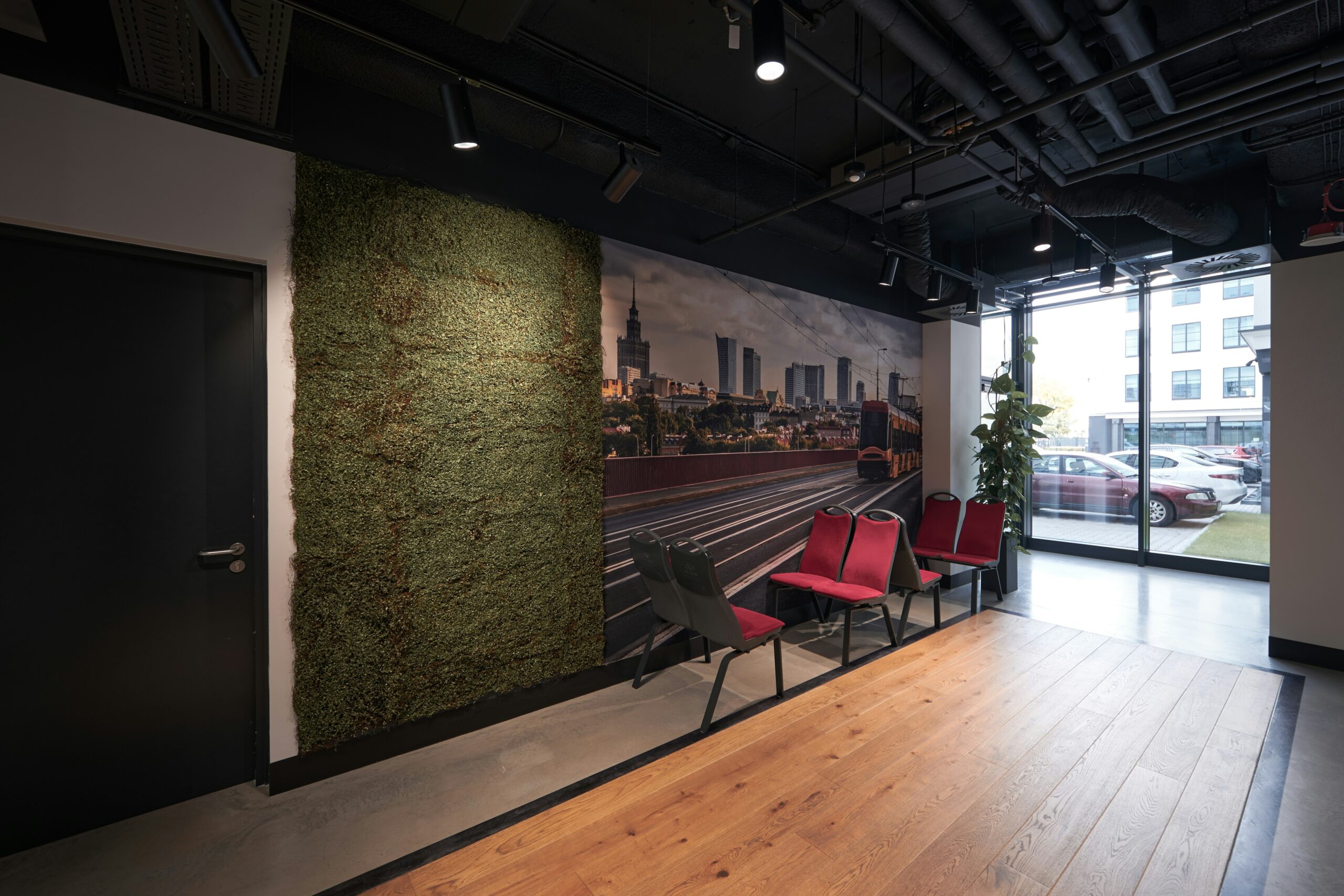 Modern office lobby with a row of red chairs, a wall covered in greenery, a cityscape mural, and large windows showing parked cars outside.