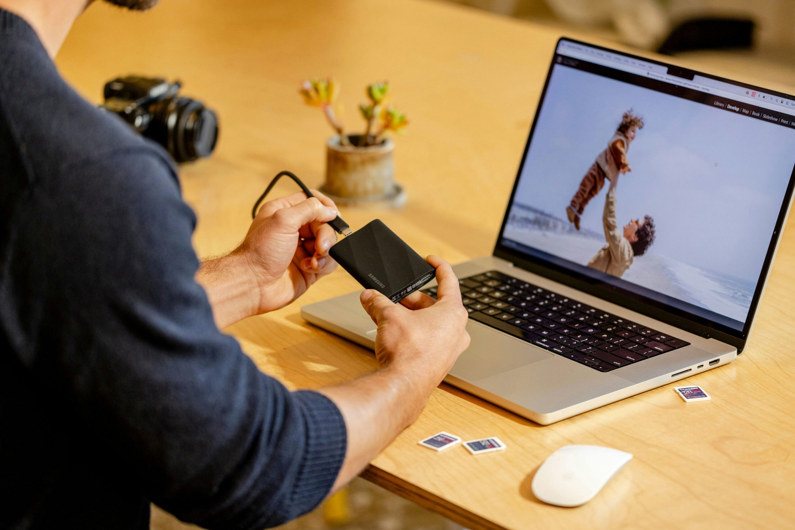 Person connecting an external hard drive to a laptop displaying a beach photo; camera, SD cards, and a plant are on the wooden table.