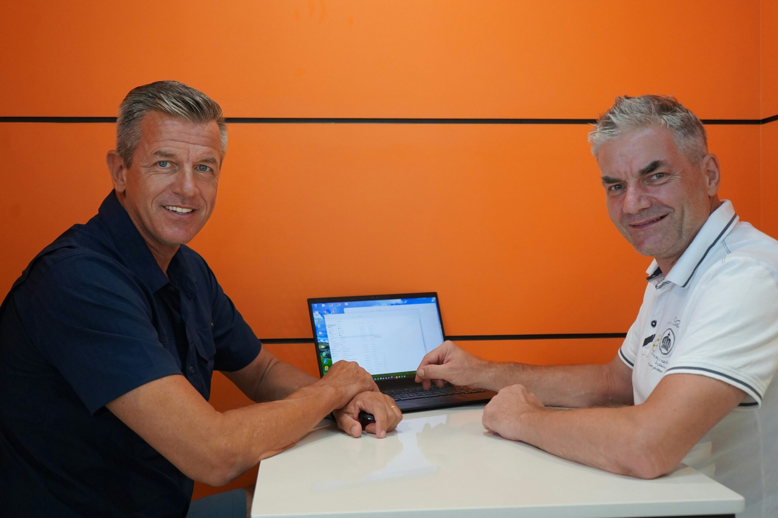 Two men sit at a table in front of an orange wall, smiling at the camera, with a laptop displaying a graph open between them.