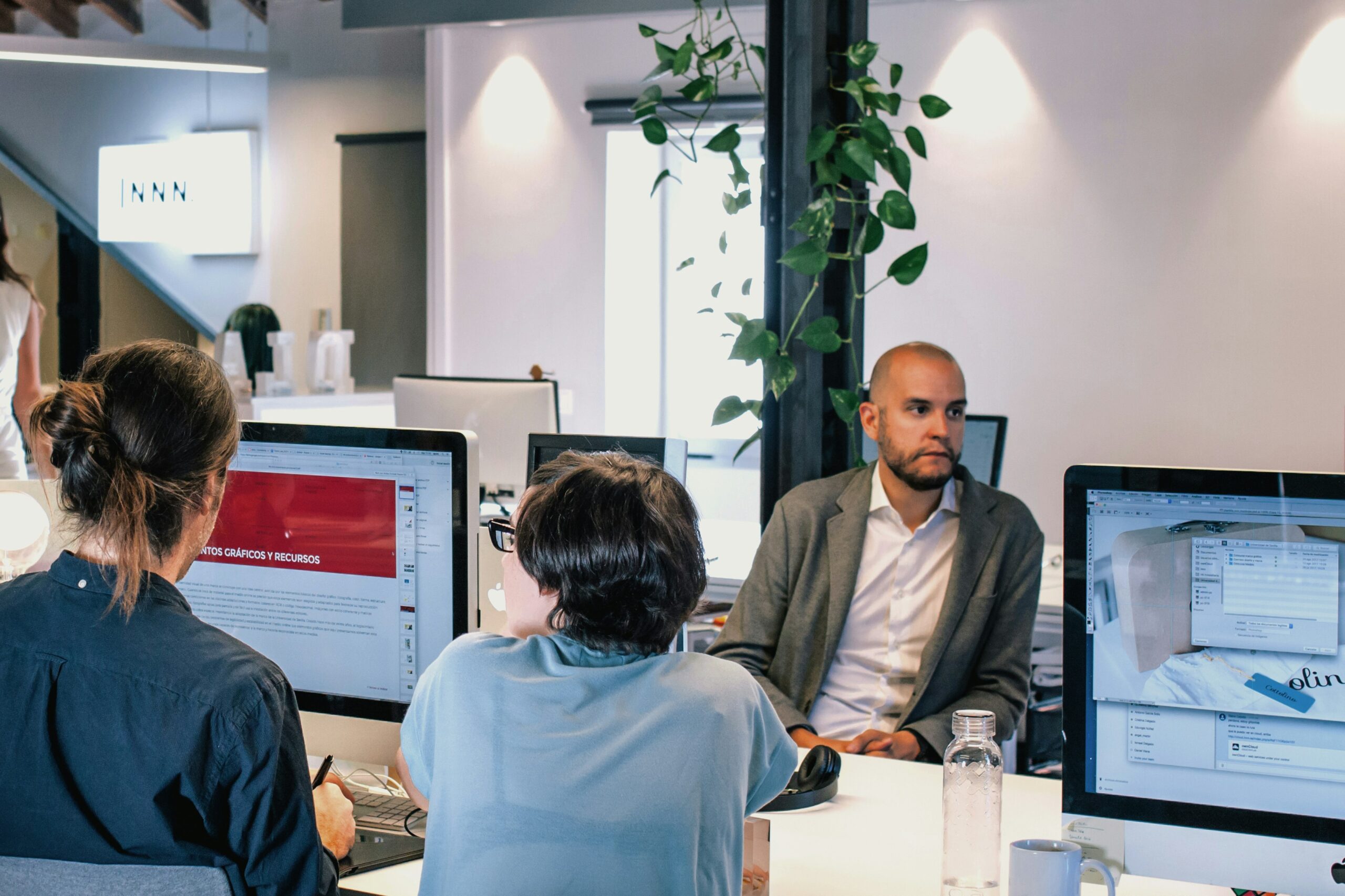 Four people are in a modern office; three are working at computers and one, wearing a suit, is seated and looking away from his desk.