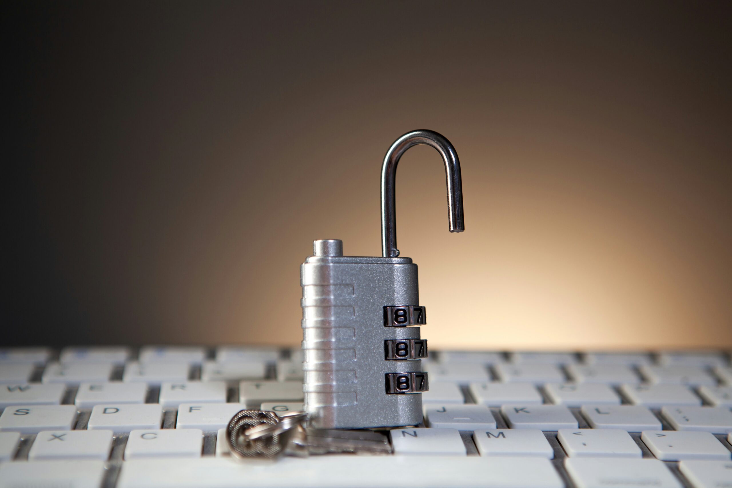 A metal combination padlock unlocked, sitting on a computer keyboard, with a set of keys placed beside it.