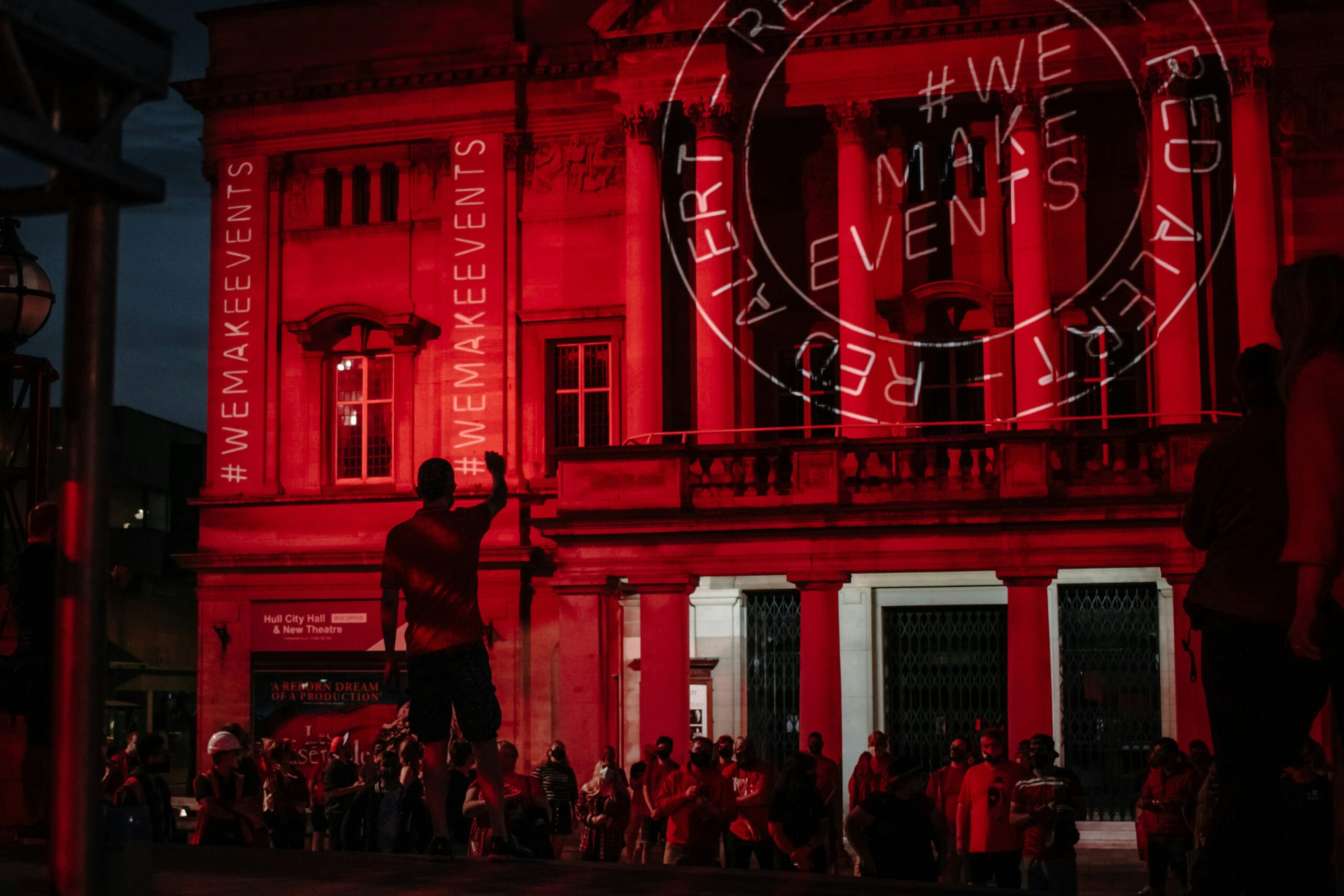 What Does a Website Maintenance Plan Actually Do? A crowd gathers outside a large building illuminated in red light, with “#WEMAKEEVENTS” and “RED ALERT” projected on its facade at night.