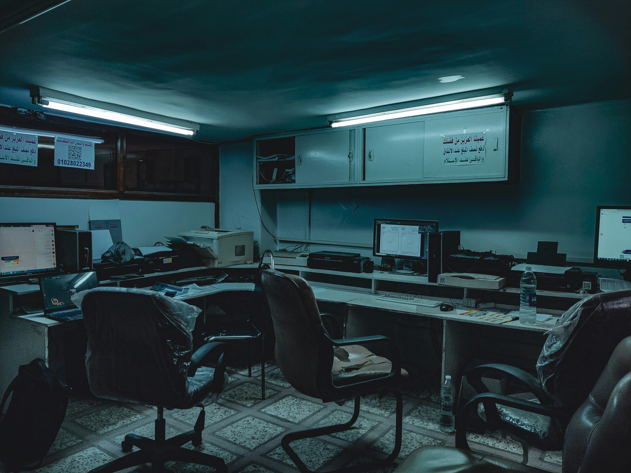protect business from malware, online safety Dimly lit office with several desks, computer monitors, chairs, and scattered office supplies under fluorescent lighting.