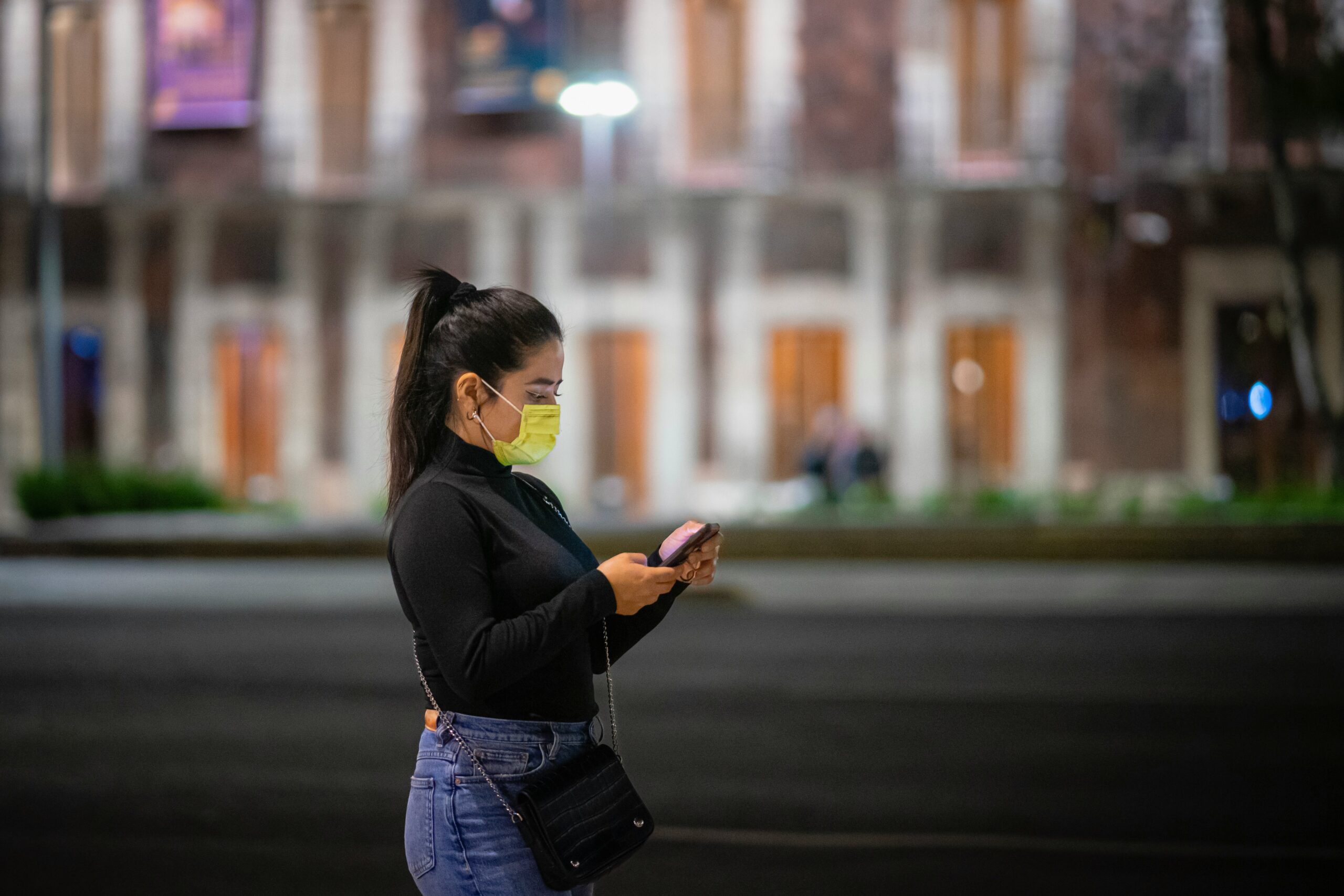 A woman wearing a yellow face mask stands on a city street at night, looking down at her smartphone.