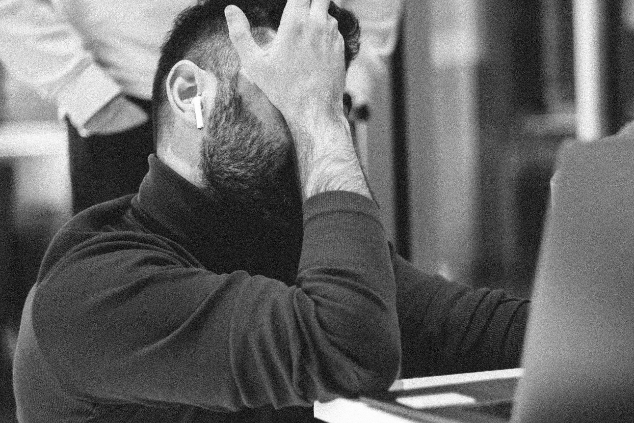 Signs Your Business Website Looks Unprofessional A man with earbuds sits at a desk with his hand covering his face, looking down at a laptop. The image is in black and white.