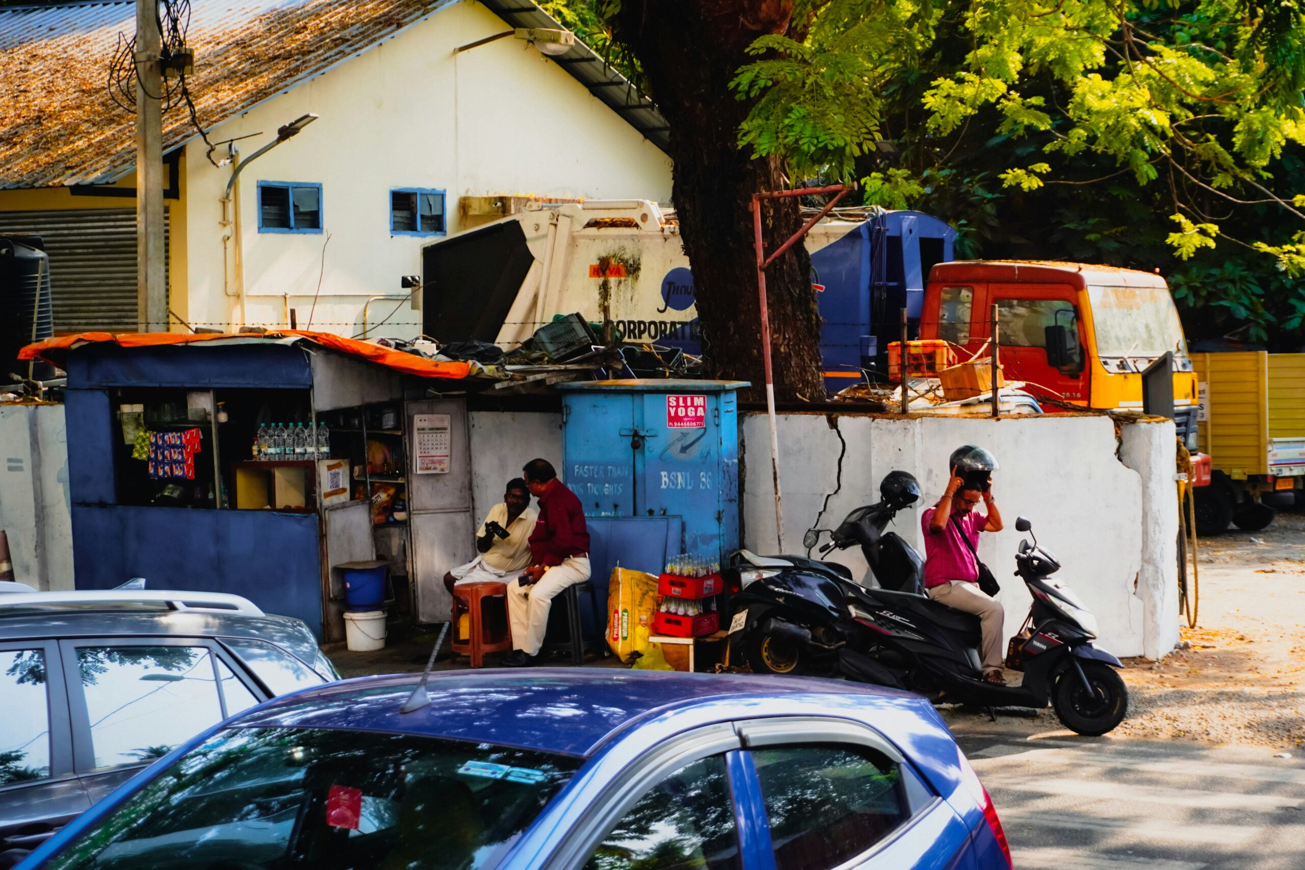 Two men in uniform are taking a break; one is sitting in a small roadside booth, while the other sits on a parked scooter. Several vehicles and a tree are nearby.