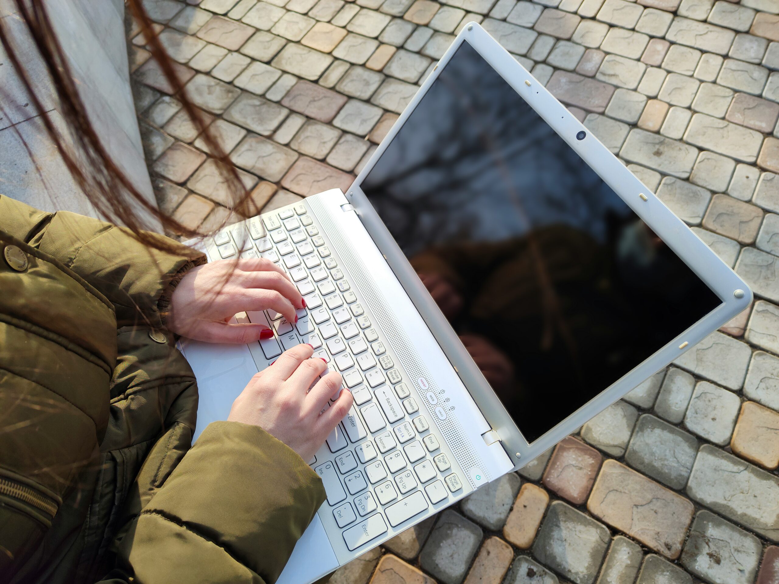 Person wearing a green jacket types on a white laptop while sitting outdoors on a stone pavement.