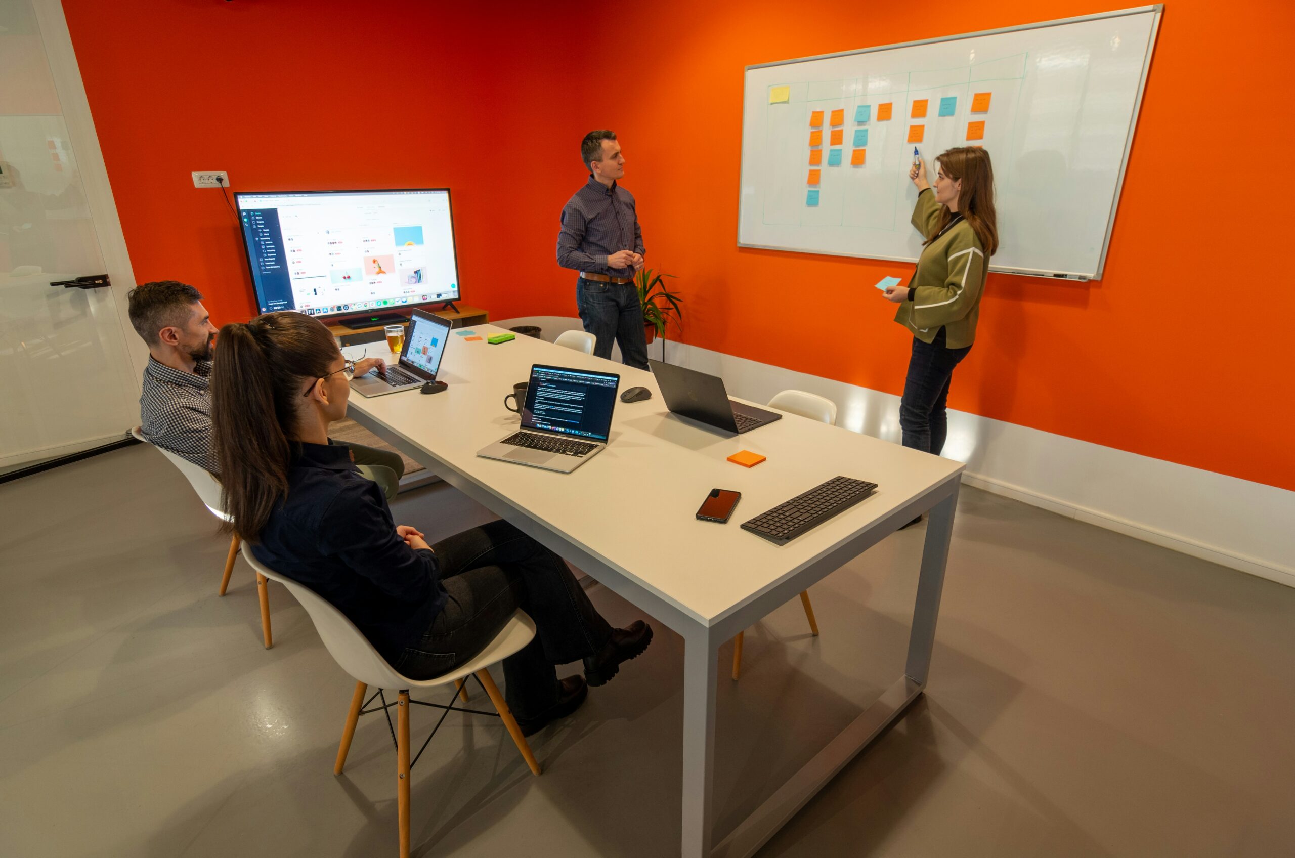Four people in a meeting room; two are seated at a table with laptops, while two stand near a whiteboard with sticky notes, discussing in front of an orange wall.