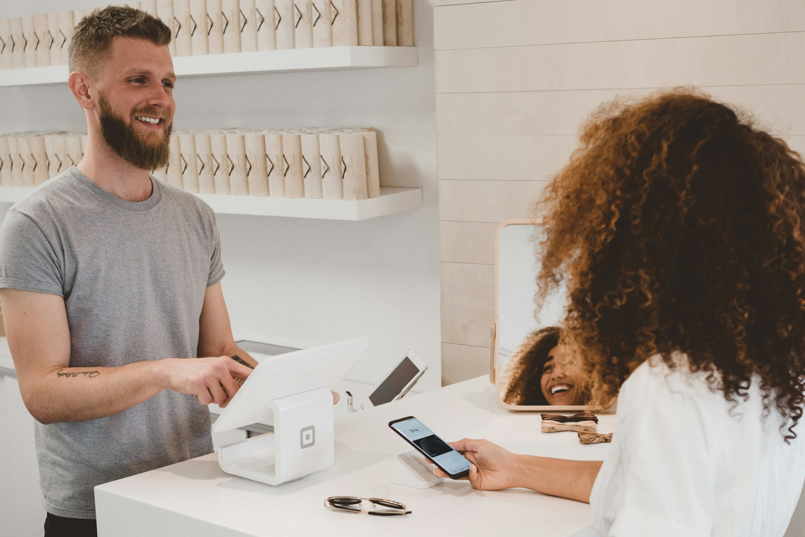 A customer uses a smartphone to pay at a store counter while a cashier stands behind the register, smiling.