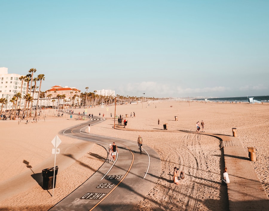 Wide sandy beach with a curving bike path, scattered people walking and cycling, palm trees, and buildings in the background under a clear blue sky.