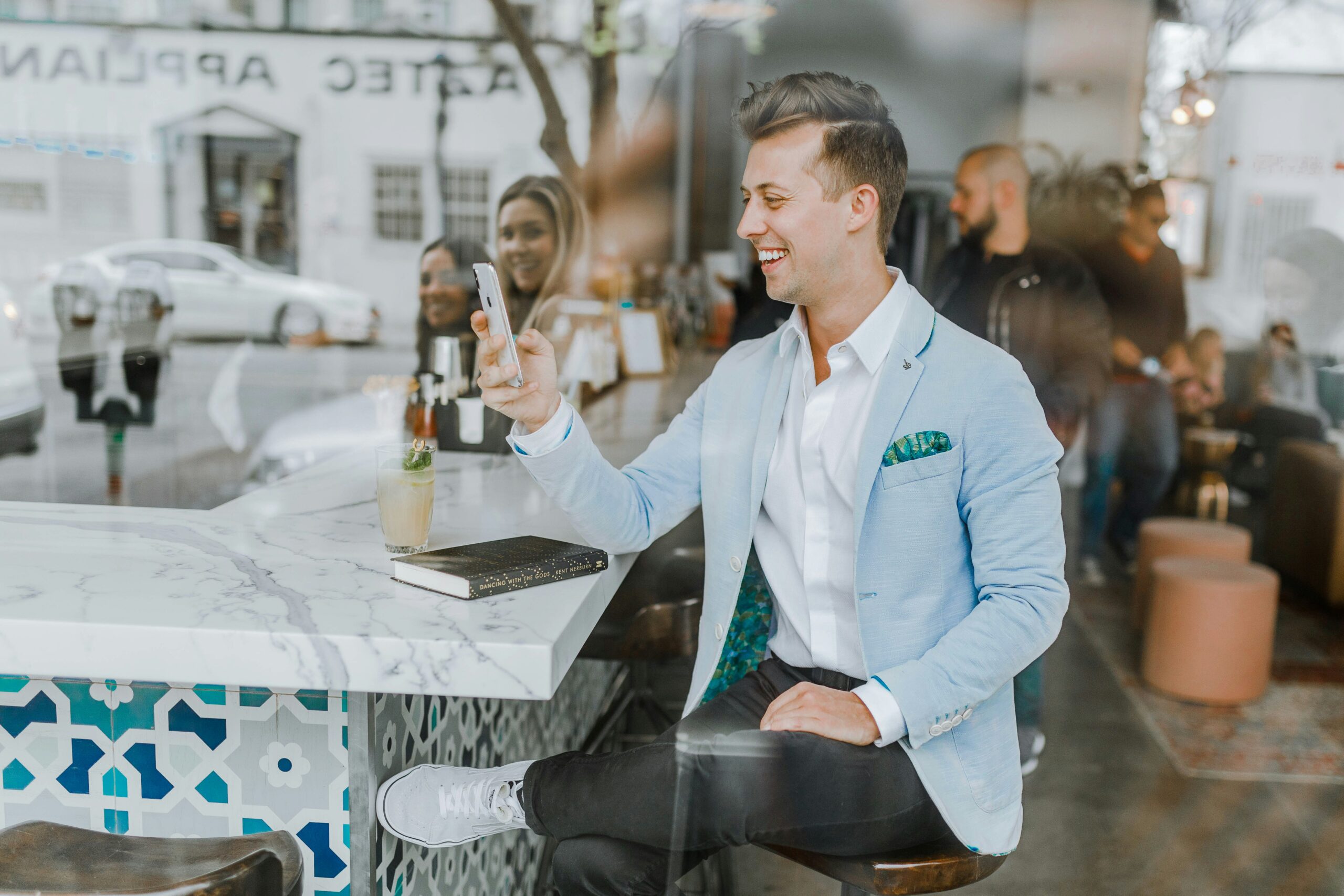 Website Backups & Maintenance: Are You Prepared for a Crash? A man in a light blue blazer sits at a marble counter in a cafe, smiling and holding a smartphone, with a drink and notebook beside him.